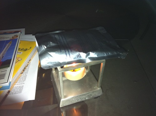 An aluminum food pouch rests on top of a cube-shaped candle holder, with a tea light candle heating the underside of the pouch.