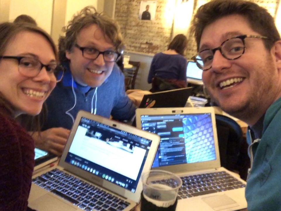 Three people crowded around laptops look into the camera, with toothy grins on their faces.