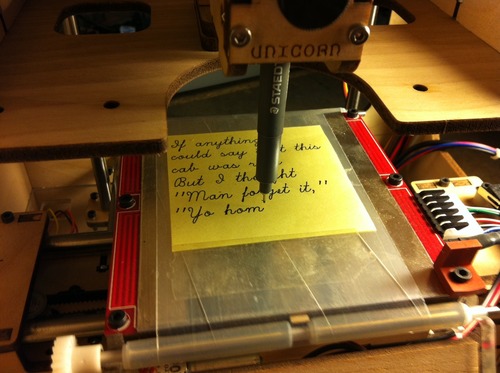 Sticky note with partially finished script text. The sticky note is on a conveyor belt platform, with a pen held in work by a Unicorn plotting tool.