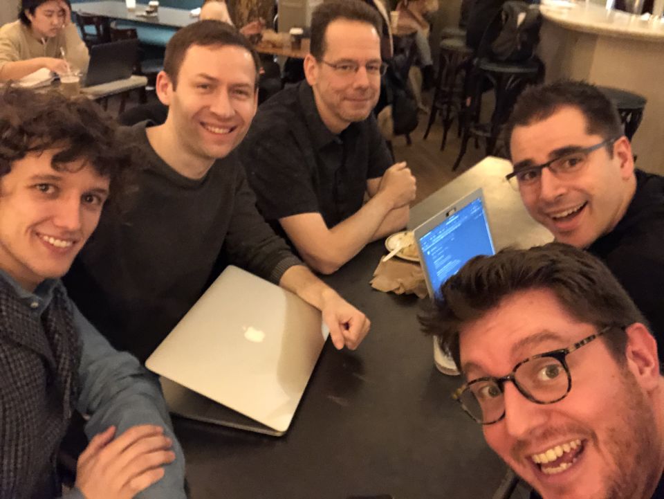 5 attendees smile at the camera, huddled around a small table with laptops on it in a busy coffee shop